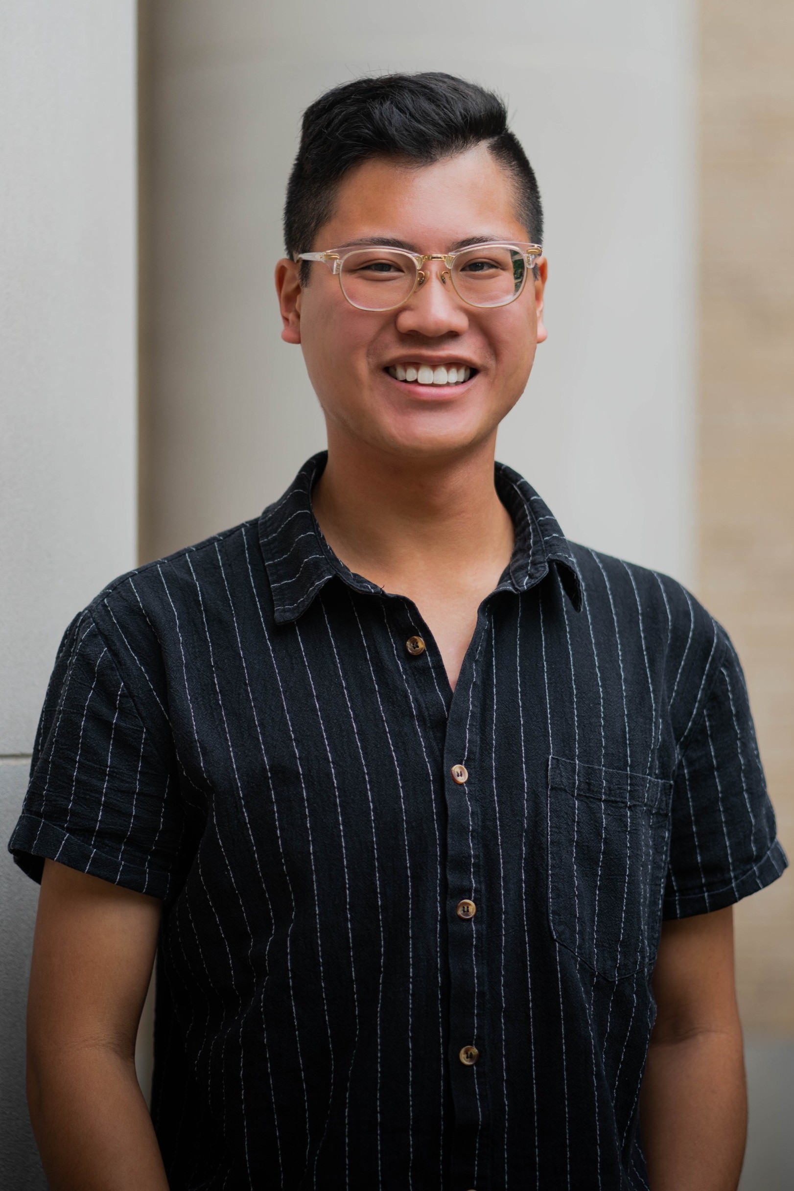 Headshot of person with short black hair and glasses, wearing a black and white stripped button down shirt.