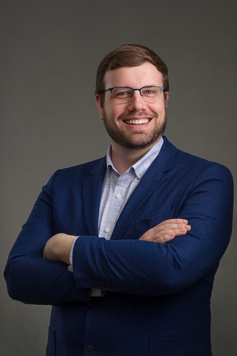 Headshot of a white person with short brown hair and glasses, in a blue patterned button-down under a blue blazer.