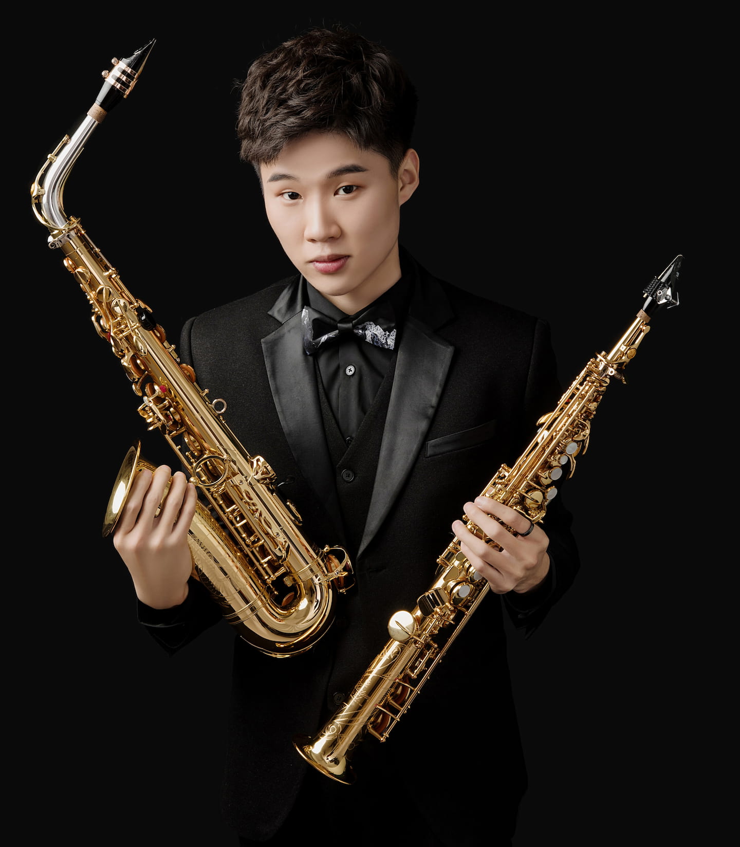 Xuanzhou Wang, dressed in a tux, holds his soprano and alto saxophones in his professional headshot
