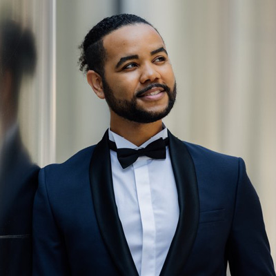 Image is of Mr. Clark leaning against a wall looking up and to his left wearing a dark colored tuxedo with a white shirt.