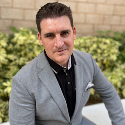 Image shows Dr. Brady seated outdoors wearing a gray blazer with a pocket square over a black shirt.