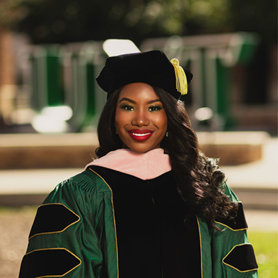 The Image shows Dr. Williams standing outdoors dressed in her regalia with three velvet black stripes trimmed in gold piping on a green gown. Her hood is draped around her neck with pink velvet. She is wearing a tam with a gold tassel.