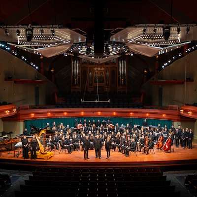 The image shows the Wind Symphony posed on the Murchison Performing Arts Center’s Winspear Performance Hall stage. The three faculty members are standing downstage. All are dressed in black.