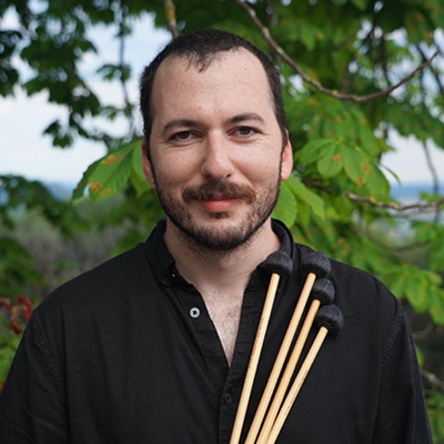 Nathan is wearing a black open collared shirt. He is holding four mallets with black heads in front of himself to his left. 