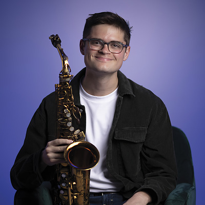 Ian is seated wearing a black coat over a white shirt holding his saxophone in front of him. There is a purple background. He wears eyeglasses with dark colored frames. 