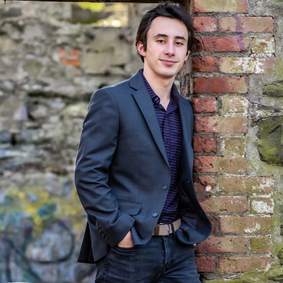 The student is leaning against a brick wall looking into the camera wearing blue jeans, a blue sportscoat over a purple striped shirt. His hands in the pants pockets.