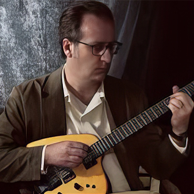 For the visually impaired: Mr. Covington is seated wearing a dark brown shirt with an open collared light colored shirt. He is playing his guitar looking down at his left hand and is wearing dark rimmed eye glasses.