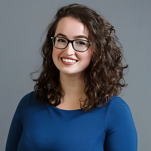 Anna Valcour, woman with shoulder length curly brown hair and glasses wearing a blue shirt.